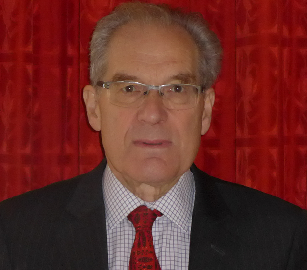Man with glasses, gray hair, suit, and red tie against a red patterned backdrop.